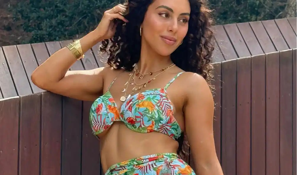 A woman with curly dark hair wearing a tropical print bikini top and bottom, adorned with multiple gold necklaces and a wide gold bracelet.