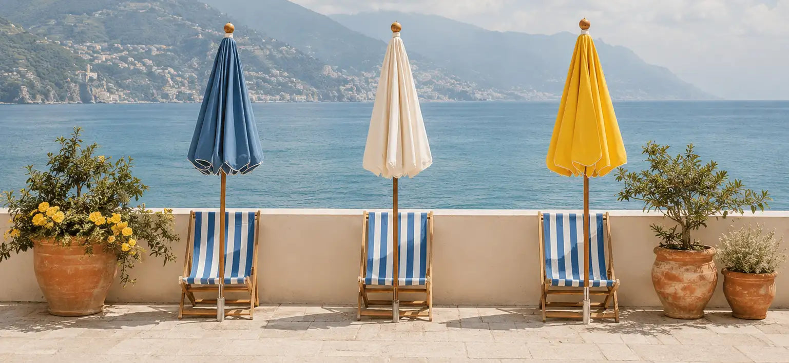 Three closed beach umbrellas in blue, cream, and yellow stand beside striped lounge chairs on a terrace overlooking the sea.