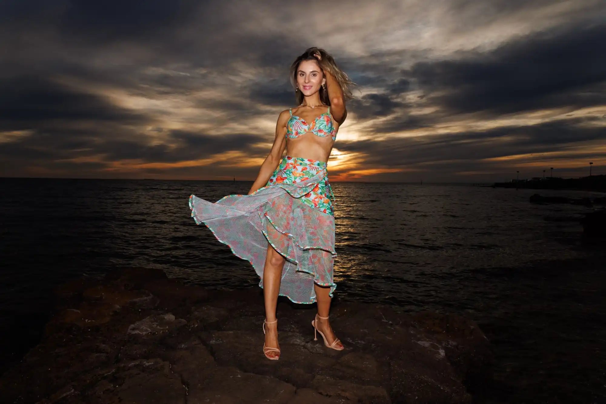 A young woman posing in a two-piece floral swimsuit with a flowing sheer skirt.